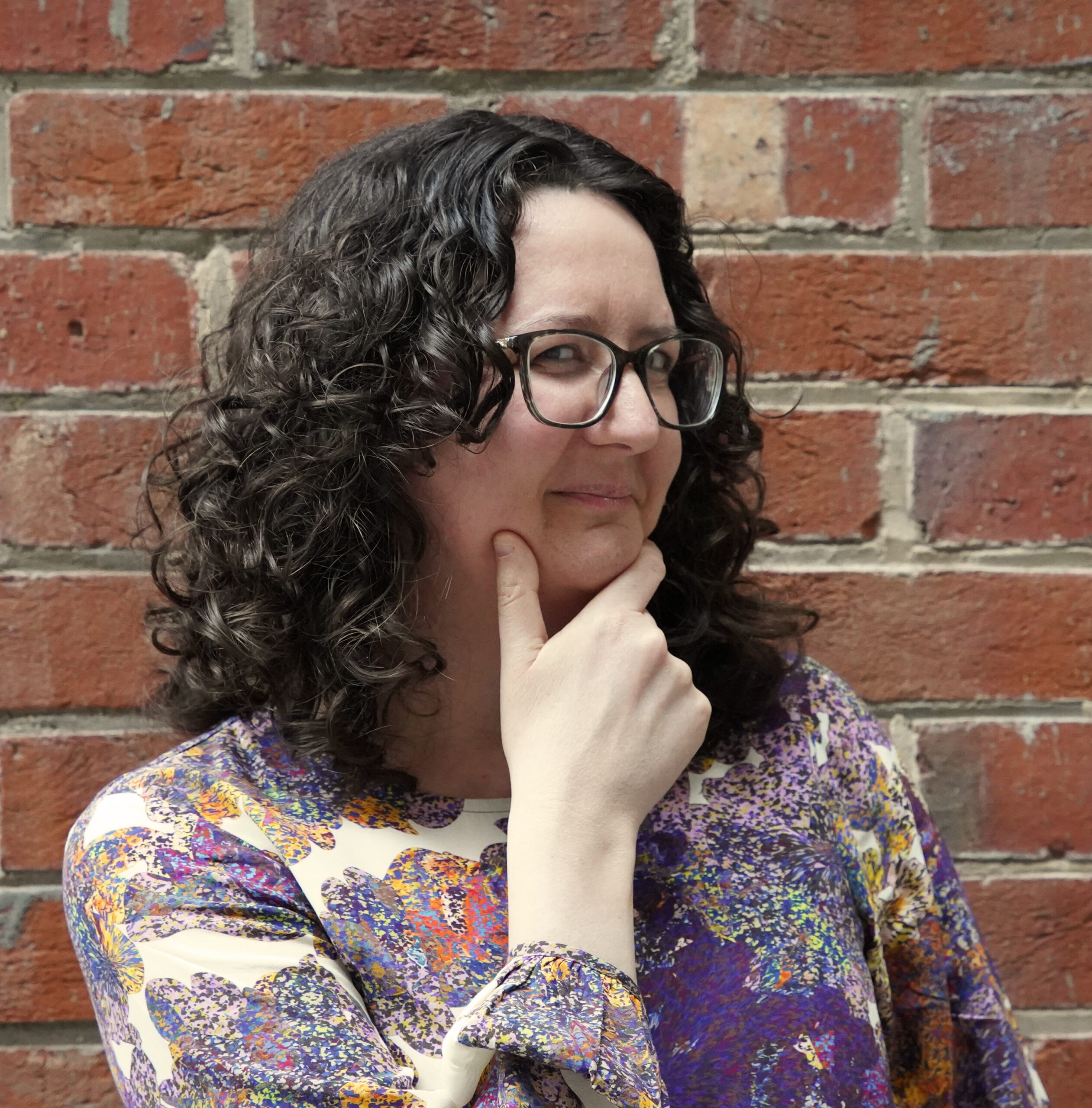 Image of the author of this website. She is a white woman in her late thirties, with dark brown curly hair and glasses. She is posing with her hand on her chin in a quizzical position. She is wearing a colourful purply-orange top.